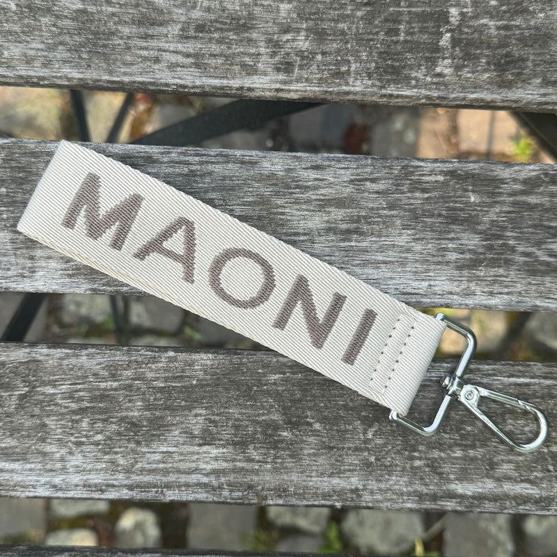 Schlichter MAONI-Schlüsselanhänger in Dunkeltaupe-Beige mit Logo-Schriftzug – dekorativ auf Holztisch arrangiert