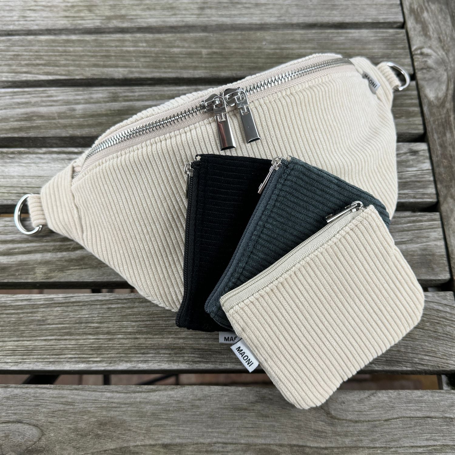 Beige Cord-Bauchtasche, hinter Etuis in dunkelgrau, beige und schwarz auf einem Holztisch präsentiert.