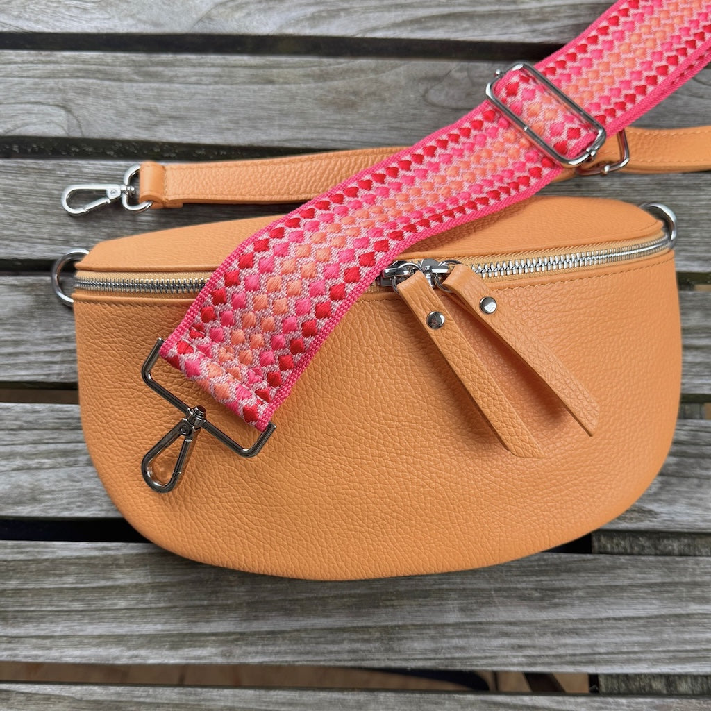 Taschen-Set auf Holztisch präsentiert: Pfirsichfarbene Bauchtasche mit schmalem Ledergurt und breitem gemustertem Gurt in neon Farben