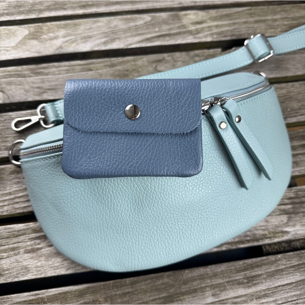 hellblaue MAONI-Bauchtasche liegt auf Holztisch, darauf liegt eine blaue Geldbörse aus Leder