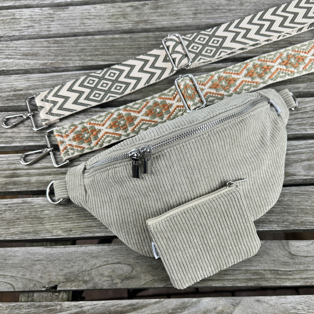 Grüne Cord Bauchtasche mit gleichfarbigen Cord Etui und zwei unterschiedlichen Wechselgurten. Die Wechselgurte liegen über der Tasche auf einem Holztisch.