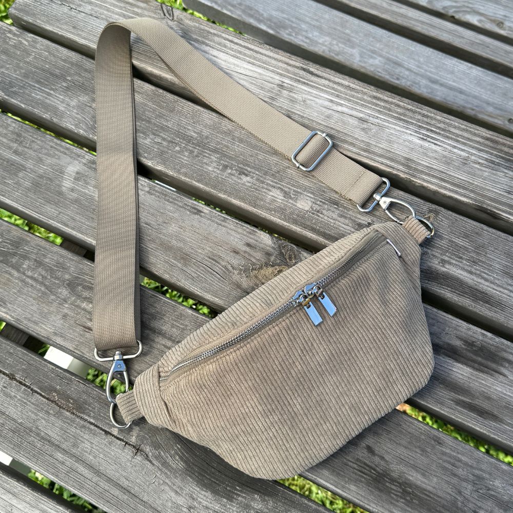 Cord Bauchtasche in der Farbe grün liegt auf einem Holz Tisch. Der Taschengurt ist verstellbar bis 140 cm und geeignet für jeden.