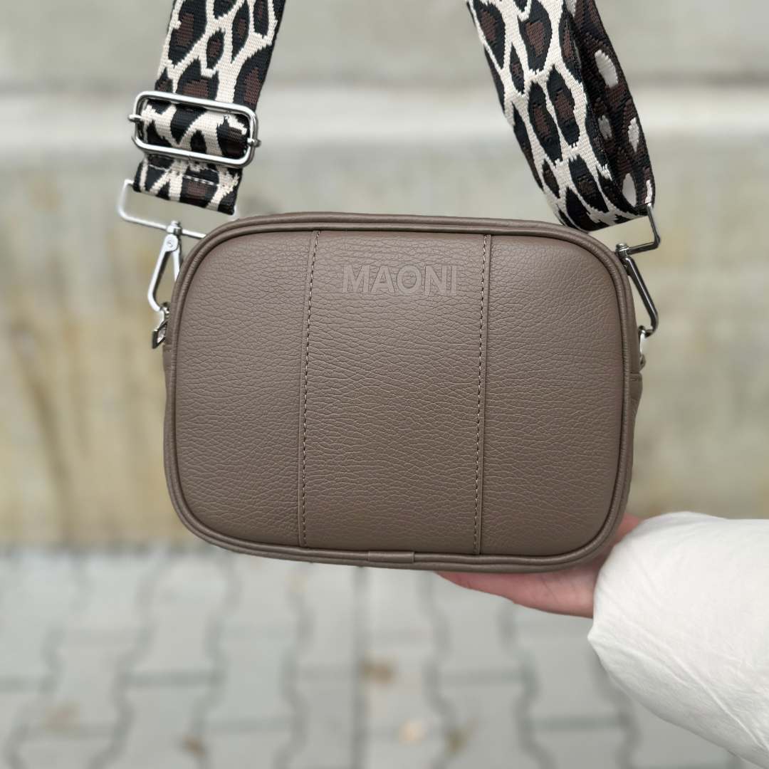 Person hält die dunkeltaupe MAONI Crossbody-Tasche mit einem breiten Leo-Gurt – ein trendiger Hingucker.