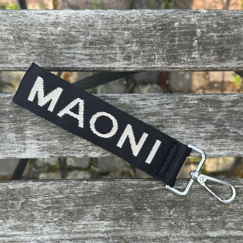 Zweifarbiger Schlüsselanhänger in Schwarz und Beige mit sichtbarem MAONI-Schriftzug – flach auf Holztisch liegend