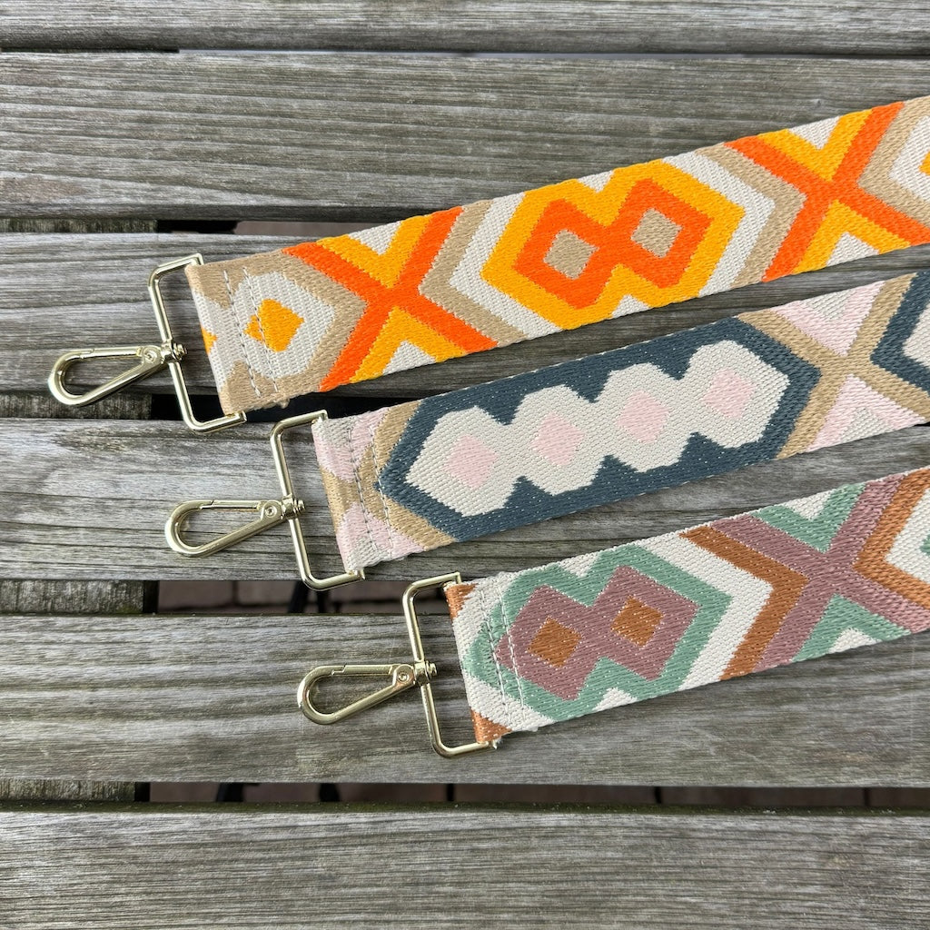 Drei Taschengurte mit bunten Mustern liegen auf einem Holztisch. Die Gurte sind in der Länge verstellbar und in den folgenden Farbgebungen: Orange-Taupe, Grau-Rosa, Rost-Grün