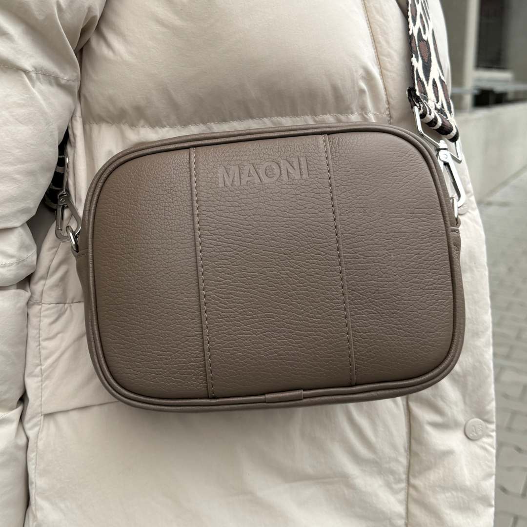 Person trägt die dunkeltaupe MAONI Crossbody-Tasche mit einem breiten Leo-Gurt – ein stylischer Hingucker.