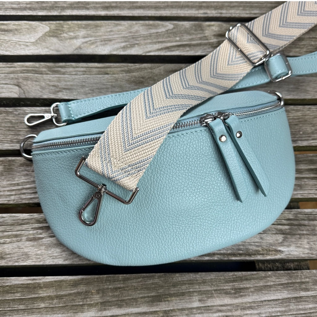 Hellblaue Bauchtasche aus Leder mit schmalem Leder- und breitem Muster-Gurt – flachliegend auf Holztisch