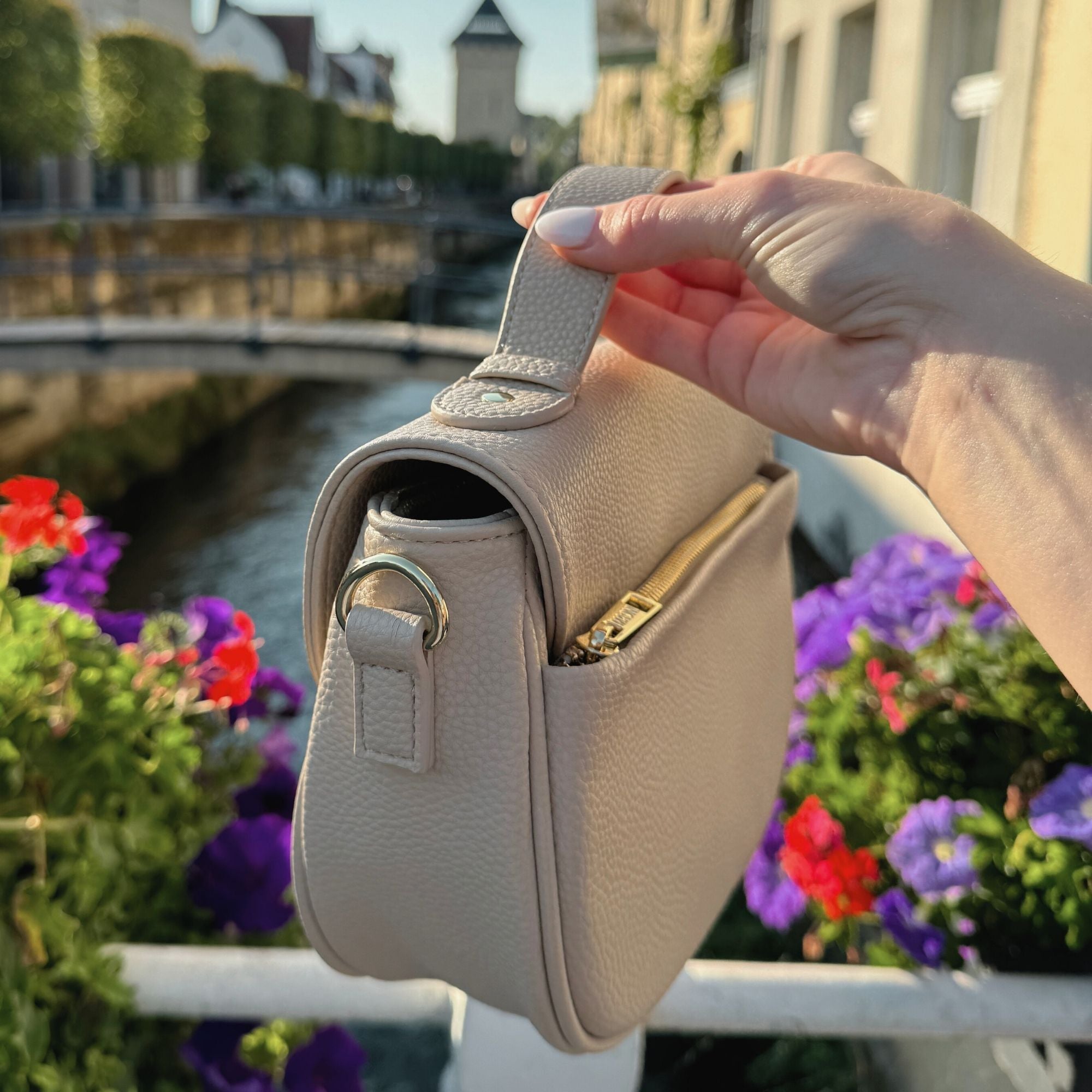Halfmoon Tasche von MAONI getragen am kleinen Henkel in einer Innenstadt mit wunderschönen roten und lilafarbenen Blumen und Fluss.
