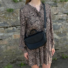 Frau trägt schwarze Halfmoon-Tasche, kombiniert mit einem Leo-Kleid, vor einer Steinwand im Hintergrund.