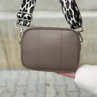 Person hält die dunkeltaupe MAONI Crossbody-Tasche mit einem breiten Leo-Gurt – ein trendiger Hingucker.